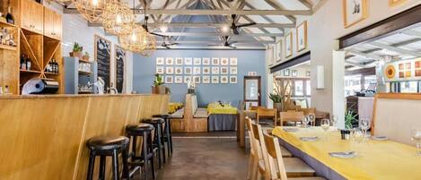 A restaurant interior with wooden bar counters, black stools, yellow tablecloths, and ceiling fans.