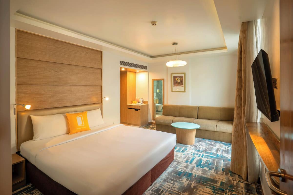 Habitación de hotel con una cama grande, un sofá, una mesita, un televisor y un baño a la vista a través de una puerta abierta.