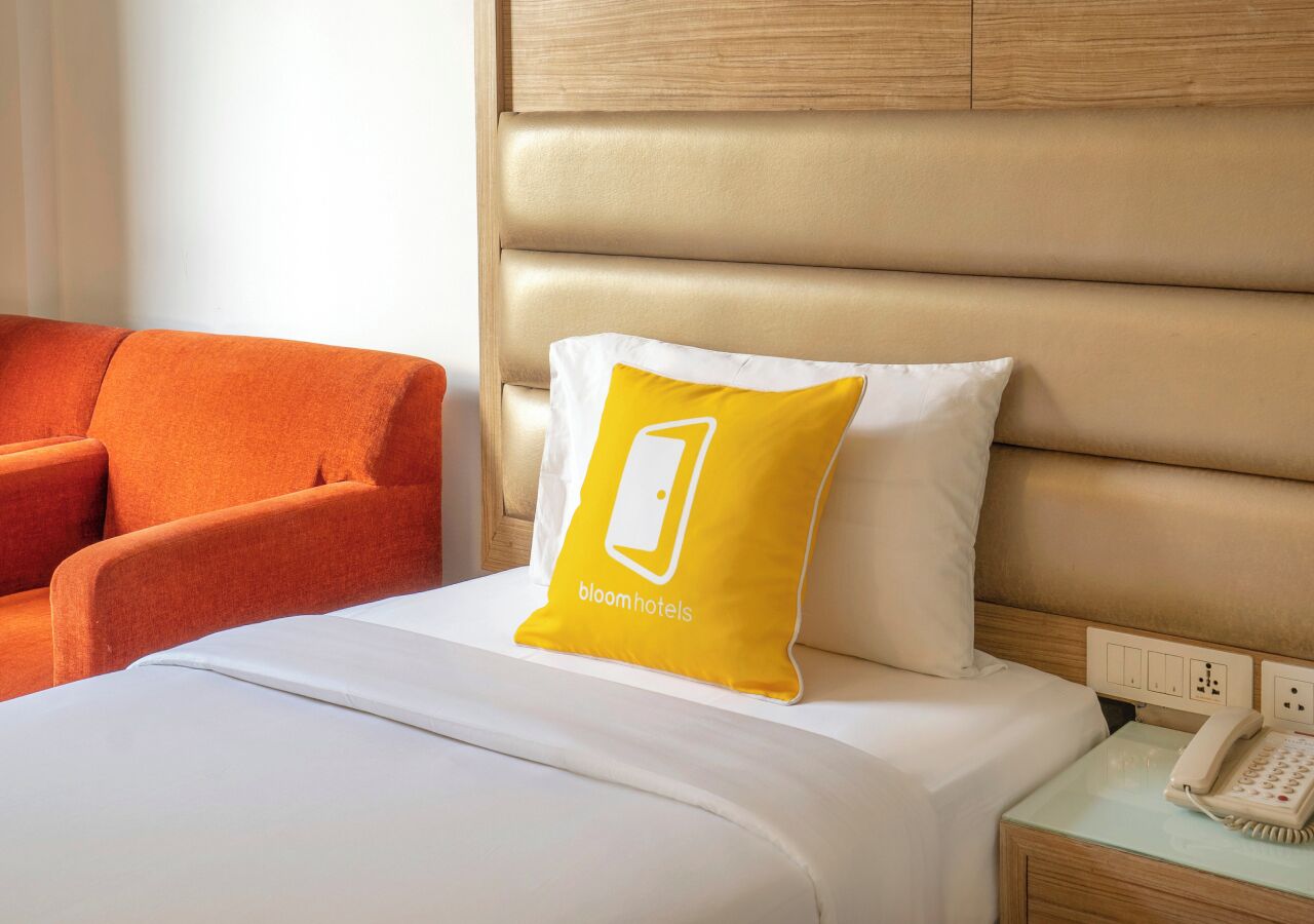 A hotel room with a bed, a bedside table with a telephone, a chair, and a pillow with a logo.