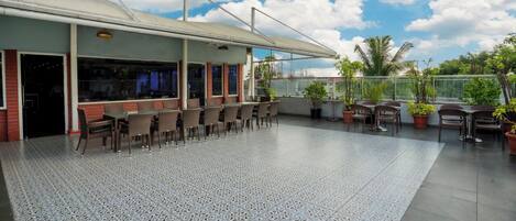 A covered outdoor seating area with tables and chairs, a tiled floor, and potted plants.