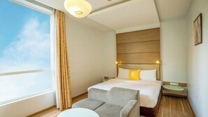 Quarto de hotel com cama, sofá, mesa pequena, mesa de cabeceira e janela com cortinas.