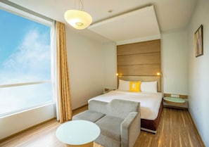 A hotel room with a bed, a sofa, a small table, a nightstand, and a window with curtains. - Bloom Boutique | Bangalore Palace Rd. (Bengaluru)