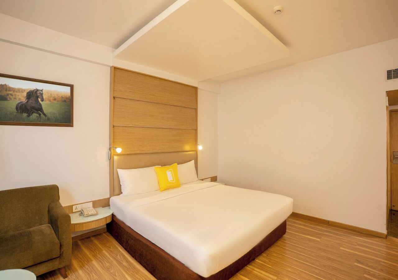 A hotel room with a bed, a nightstand, a chair, a painting, and a wooden headboard.