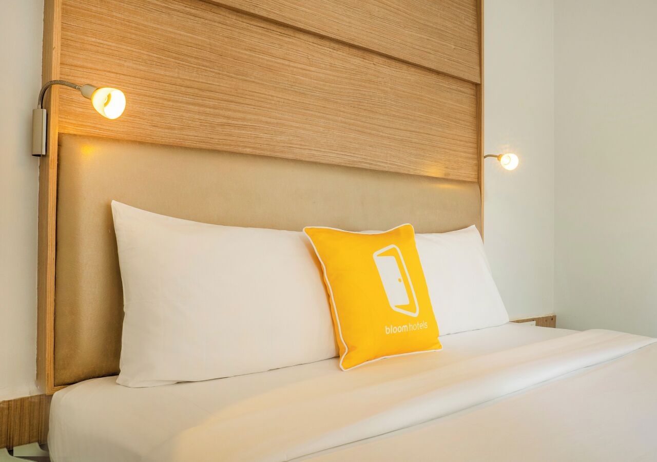 Habitación de hotel con cabecera de madera, ropa de cama blanca y almohada amarilla con un logo.