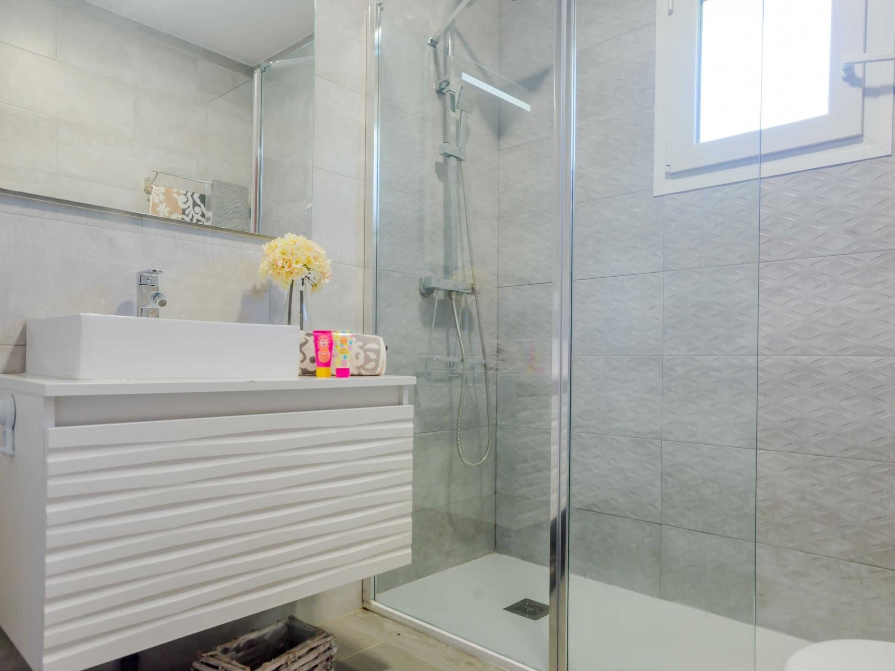 A modern bathroom with a glass shower enclosure, a white vanity with a mirror, and a small vase of flowers.