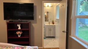 A bedroom with a TV, a wooden dresser, a floral bedspread, and a view of a bathroom with a mirror, sink, and window. - Enjoyable 3-bedroom house in charming Surrey neighborhood, close to everything (Surrey)