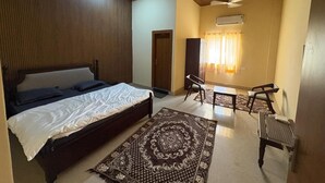 A bedroom with a bed, a desk, two chairs, a patterned rug, and a ceiling fan. - Hotel Bhargav Palace (Jaisalmer)