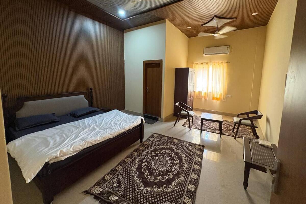 A bedroom with a bed, a desk, two chairs, a patterned rug, and a ceiling fan.