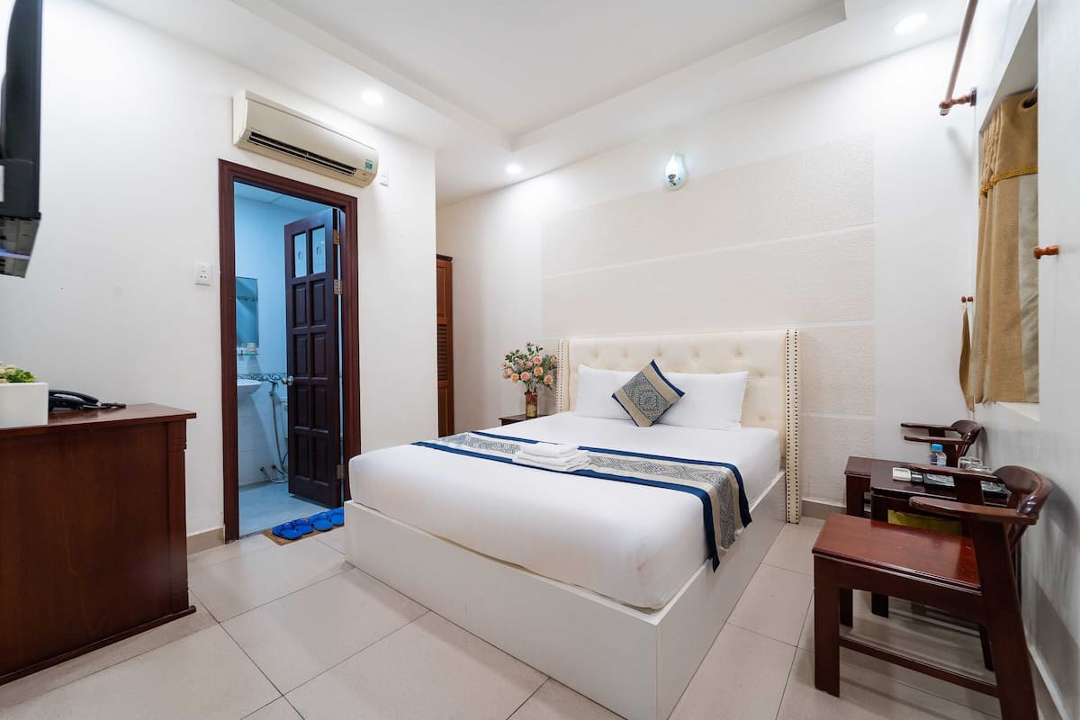 A hotel room with a bed, a desk, a chair, a mirror, and an air conditioning unit.