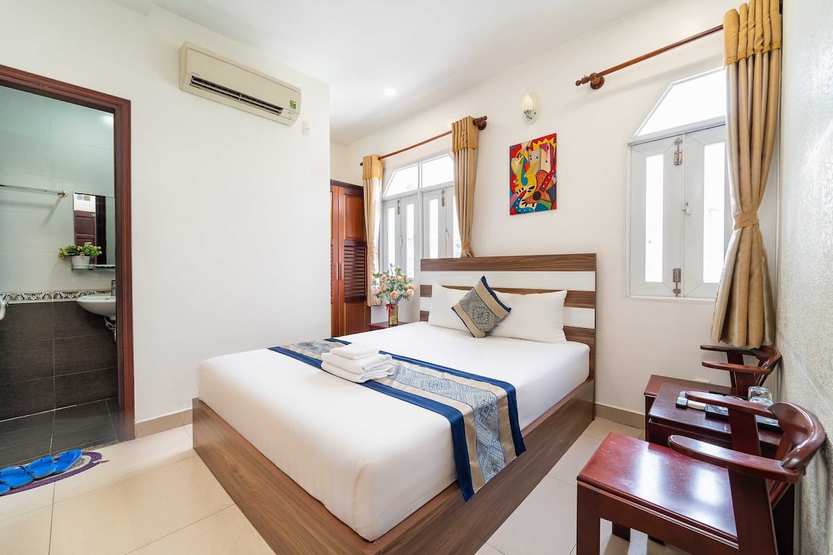 A hotel room with a bed, a desk, a chair, a window with curtains, an air conditioning unit, and a painting on the wall.