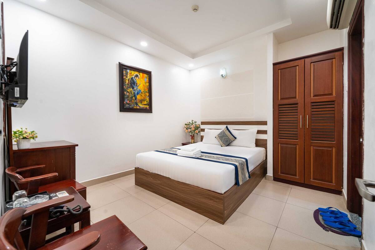 A hotel room with a bed, wooden wardrobe, desk with telephone, and a painting on the wall.