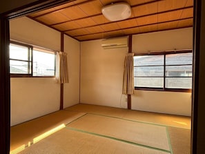 Una habitación con suelo de tatami, vigas de madera, ventilador de techo y dos ventanas con cortinas.