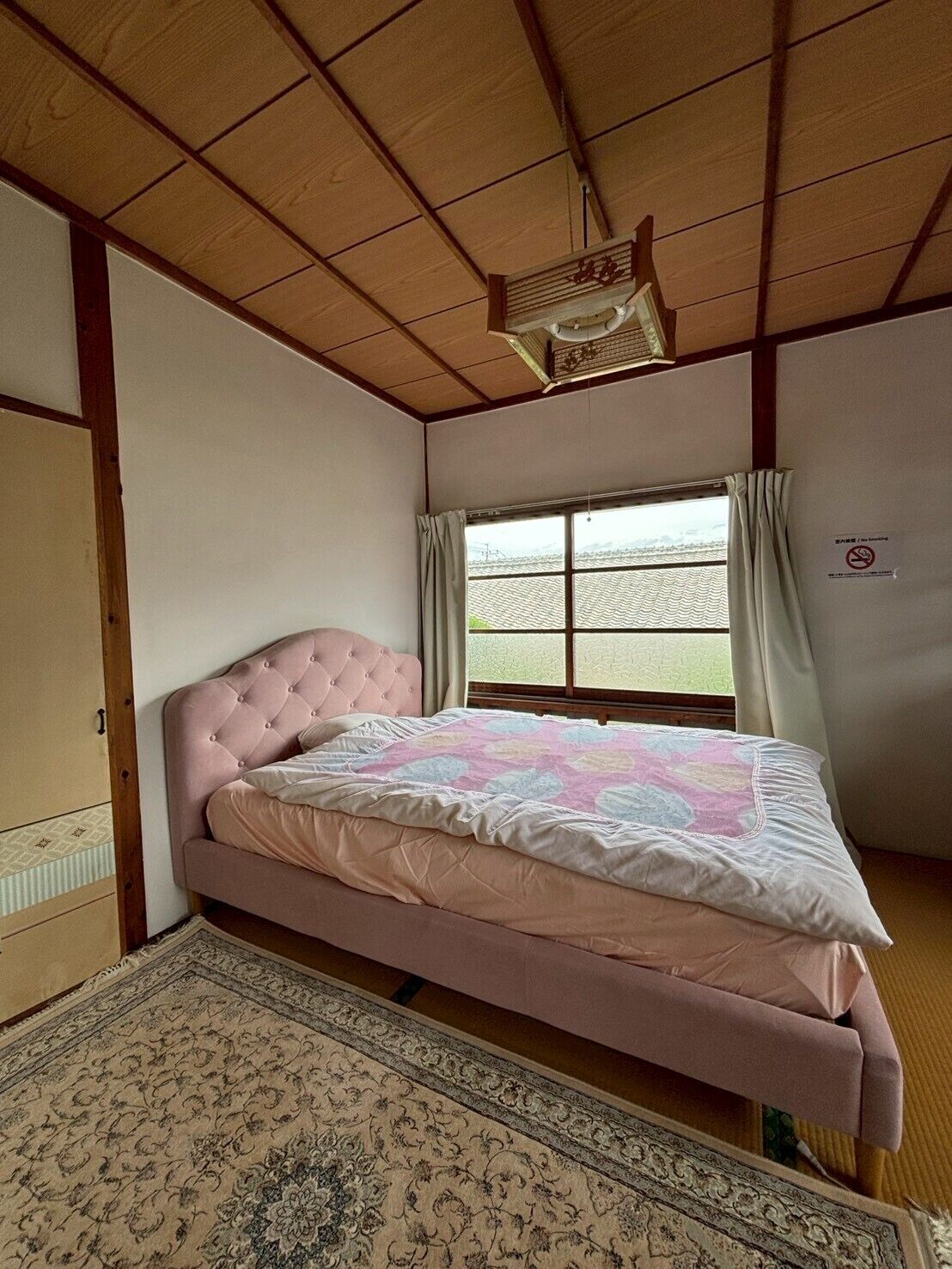Une chambre avec un lit rose, un plafond en bois, une fenêtre avec des rideaux et une porte coulissante de style japonais traditionnel.