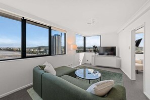 Ein modernes Wohnzimmer mit einem grünen Sofa, einem Glastisch und einem Fernseher. Große Fenster bieten Blick auf die Stadt.