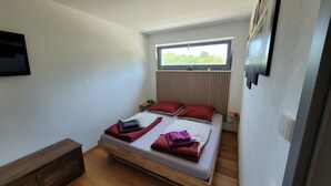 Een kleine, goed verlichte slaapkamer met een enkel bed, een houten nachtkastje, een aan de muur bevestigde televisie, en een raam met uitzicht op groen.
