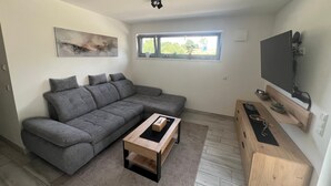 Een moderne woonkamer met een grijze hoekbank, een houten salontafel, een wandgemonteerde flatscreen televisie en een klein houten kastje.