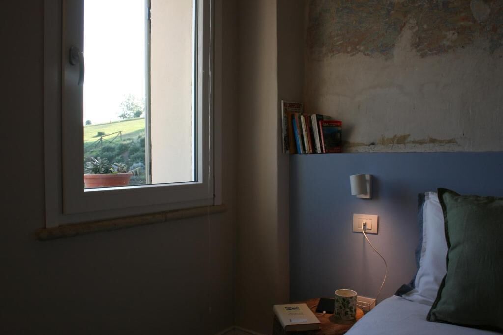 A bedroom with a window, a bedside table with a lamp, a bed with pillows, and a wall-mounted shelf with books.