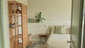 A bedroom with a bed, a wooden bookshelf, a small table, and a potted plant.