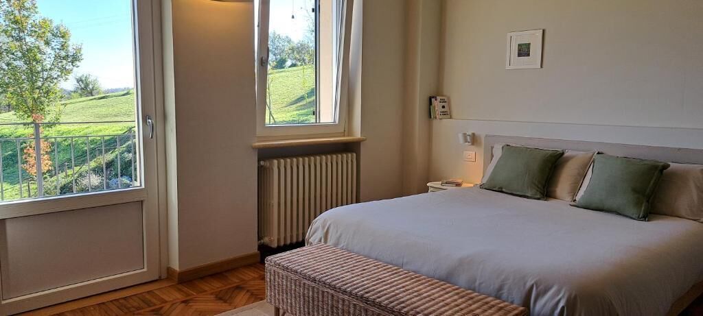 A bedroom with a large bed, a window with a view of a green landscape, a radiator, and a bedside table with a lamp.