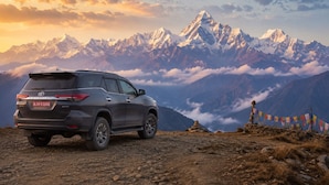A vehicle parked on a mountain road with a view of snow-capped peaks and prayer flags in the background.