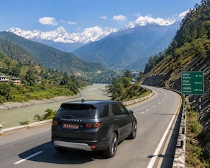 A Discovery SUV driving on a mountain road with a river on the left and a green sign indicating distances to Mugling, Pokhara, and Jomsom.