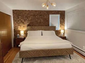 Ein Schlafzimmer mit Holz-Kopfteil, einem Bett mit weißer Bettwäsche, zwei Nachttischen mit Lampen, einem Fenster mit Jalousien und einem Holzschrank.