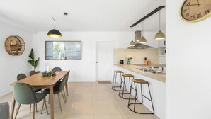 Une cuisine moderne avec un coin repas, comprenant une table en bois, des tabourets de bar, des luminaires suspendus et une grande horloge murale.