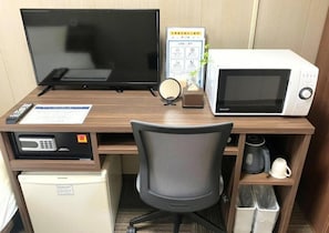 A hotel room with a wooden desk, a television, a microwave, a refrigerator, a chair, and a coffee maker.
