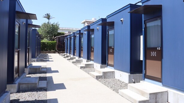 Fila de habitaciones de hotel azules con puertas numeradas, un camino de hormigón y un cielo despejado.
