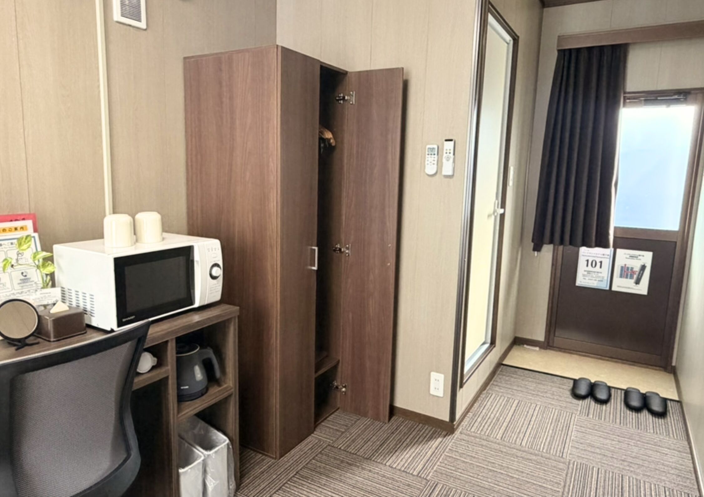 A hotel room with a microwave, a small refrigerator, a desk with a chair, a wardrobe, a door with a number sign, and a window with curtains.