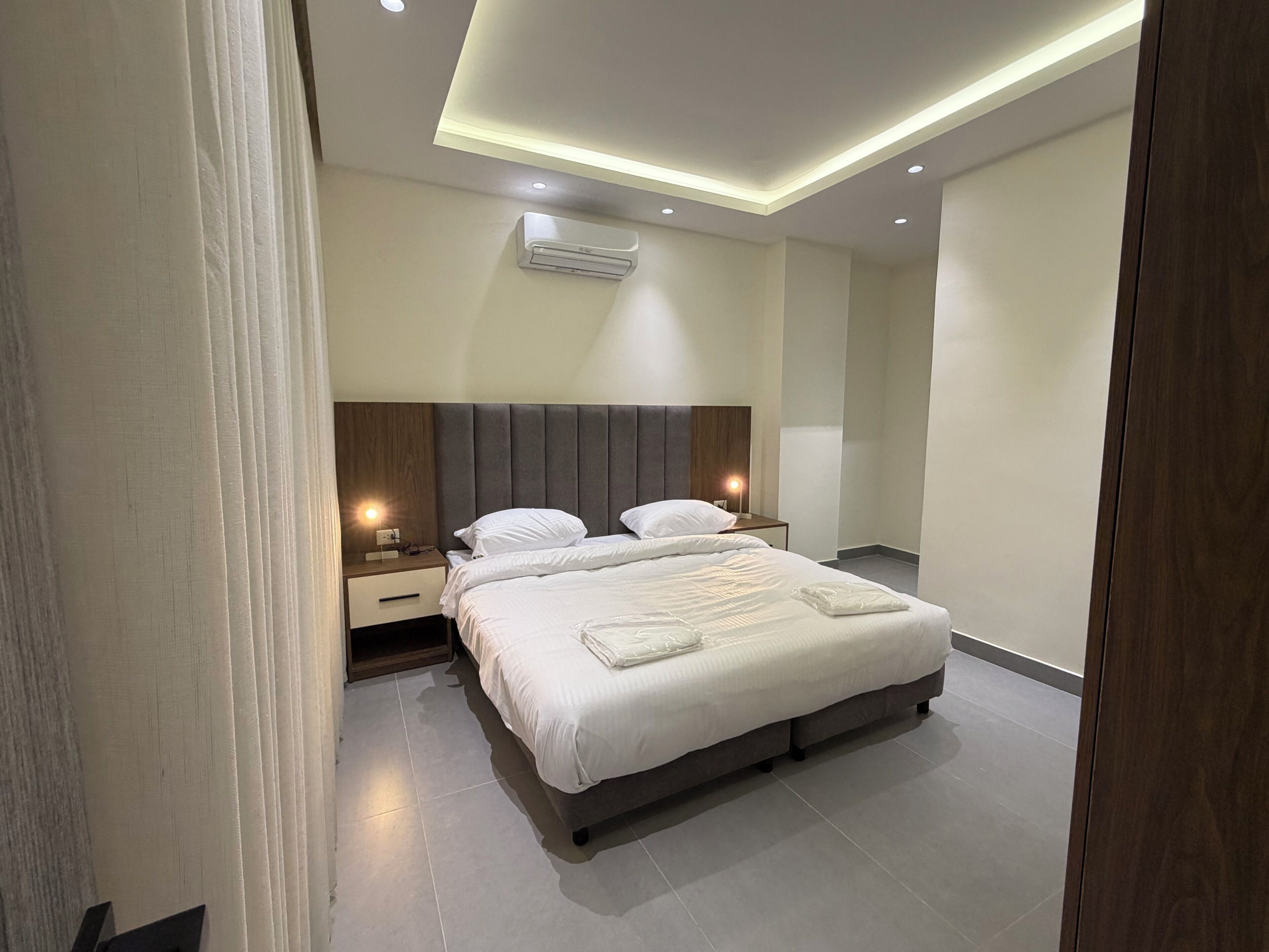 A modern hotel room with a large bed, a nightstand, a wall-mounted air conditioner, and recessed ceiling lighting.