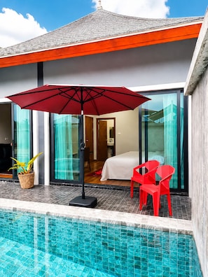 Ein modernes Hotelzimmer mit Glastür, die auf einen Balkon führt, einem roten Sonnenschirm und einem Poolbereich.