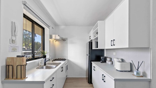 A modern kitchen with white cabinetry, a built-in stove, and a window overlooking a balcony. - 15 Calm & Cosy w Parking - Walk to Beach & Train (Mosman Park)