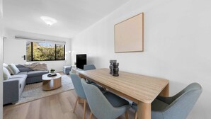 A modern living room with a wooden dining table, grey upholstered chairs, a grey sofa, a small coffee table, and a television. - 15 Calm & Cosy w Parking - Walk to Beach & Train (Mosman Park)