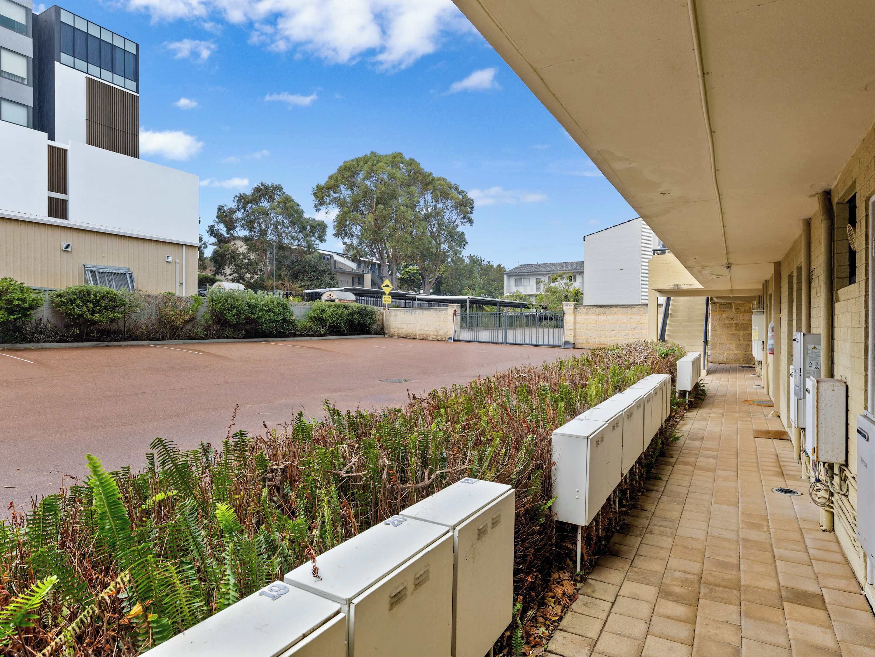 A parking area with a building on the left, a fenced area, and vegetation in the foreground.