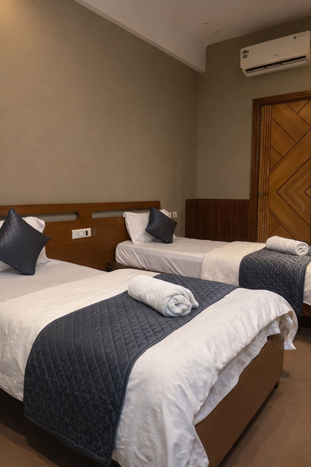 A hotel room with two beds, each with a quilted bedspread and pillows, a wooden headboard, and an air conditioning unit on the wall.
