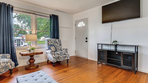 Ein Zimmer mit Holzdielenboden, einem großen Flachbildfernseher an der Wand, einem gemusterten Sessel, einem kleinen Tisch mit Pflanze und einem grauen Pouf.