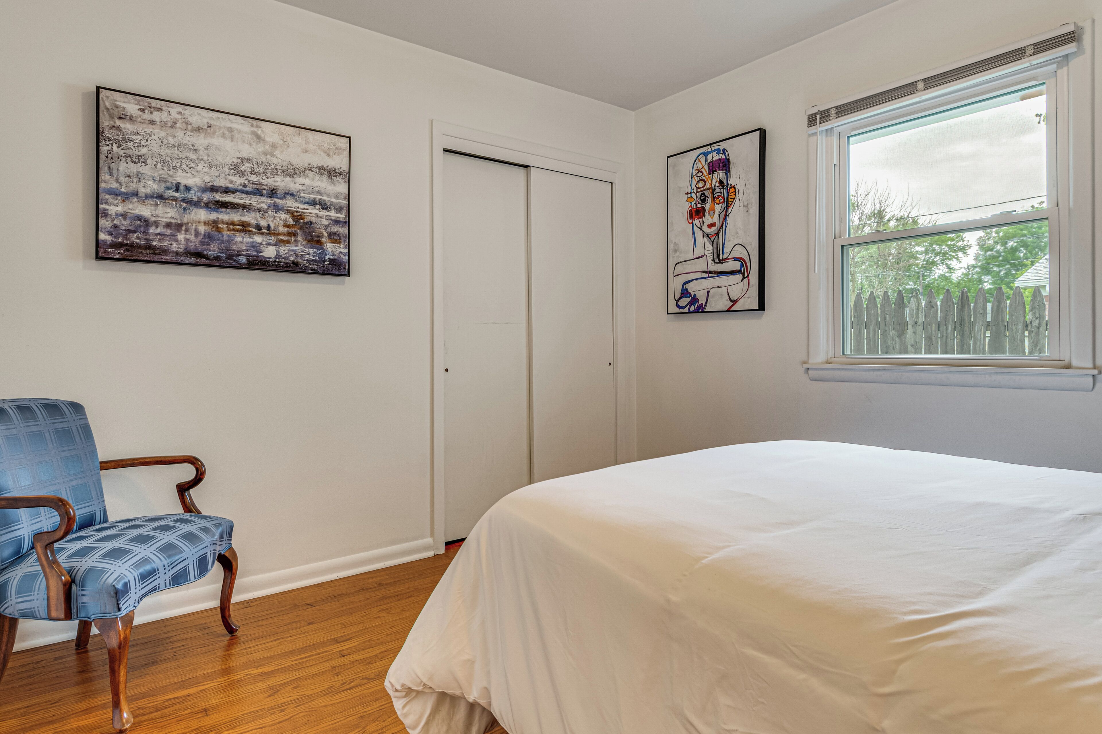 A bedroom with a bed, a chair, two paintings, a window, and a door.