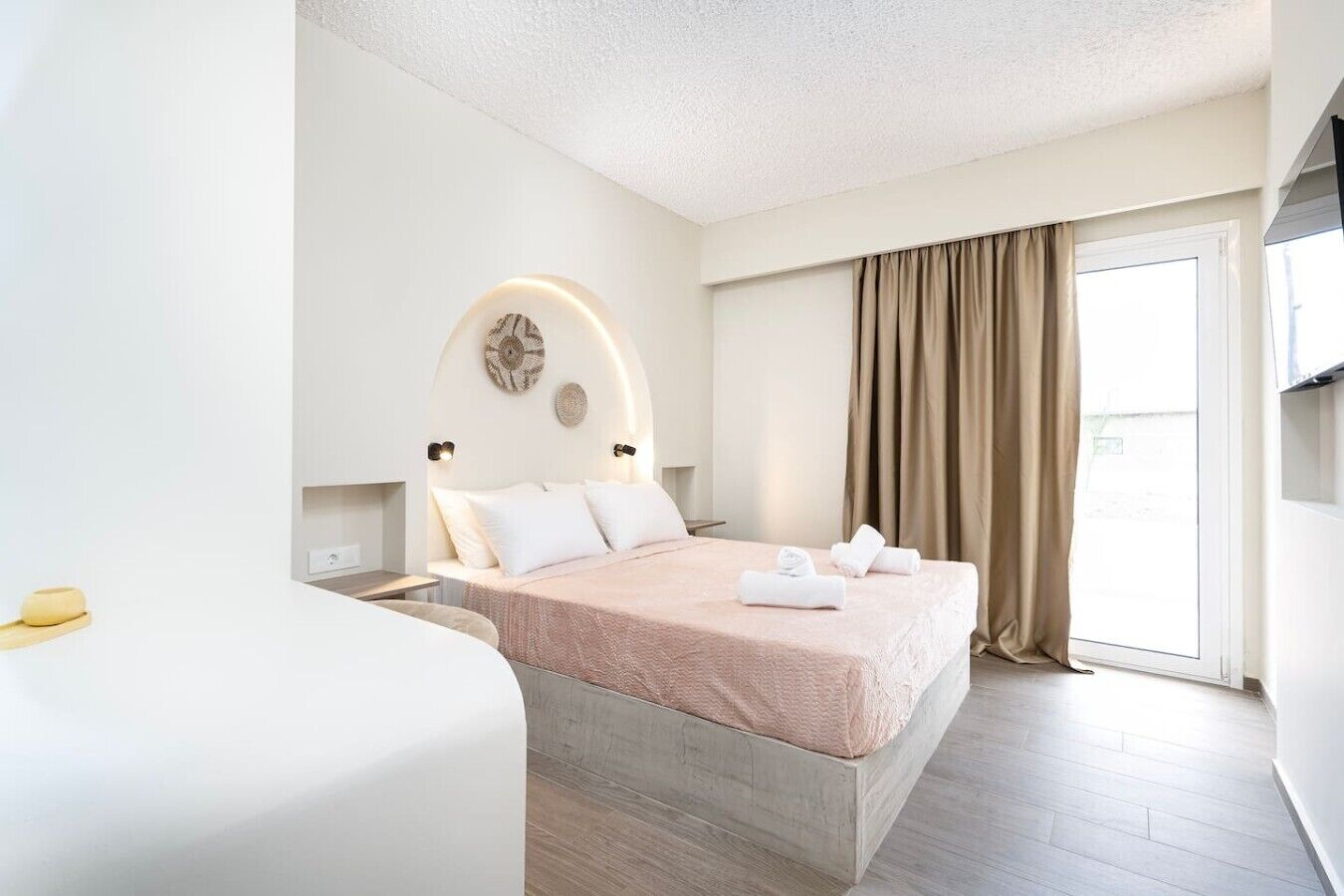 A hotel room with a bed, a bedside table, a lamp, a window with curtains, and a view outside.