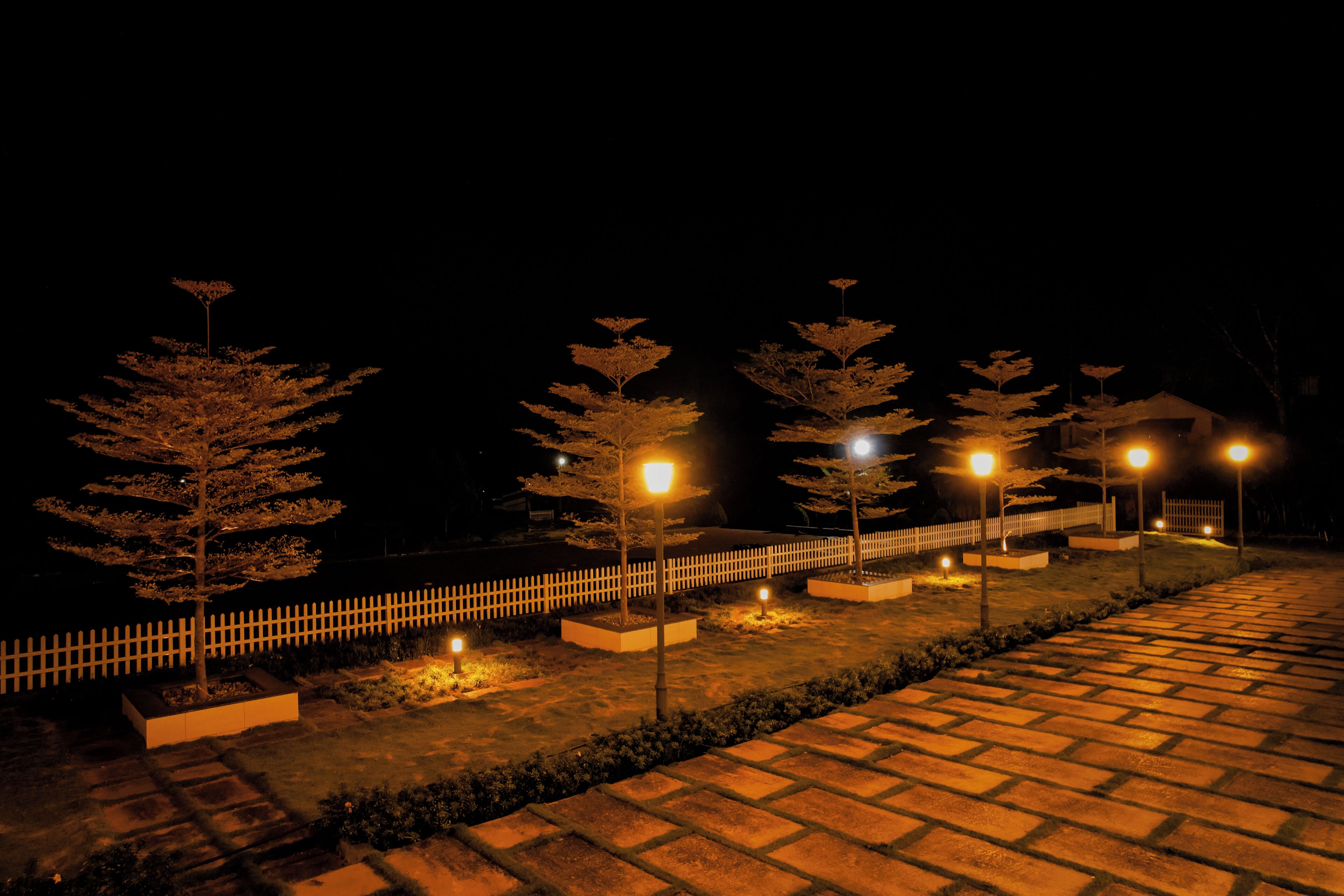 A nighttime outdoor pathway with evenly spaced street lamps and a white picket fence.