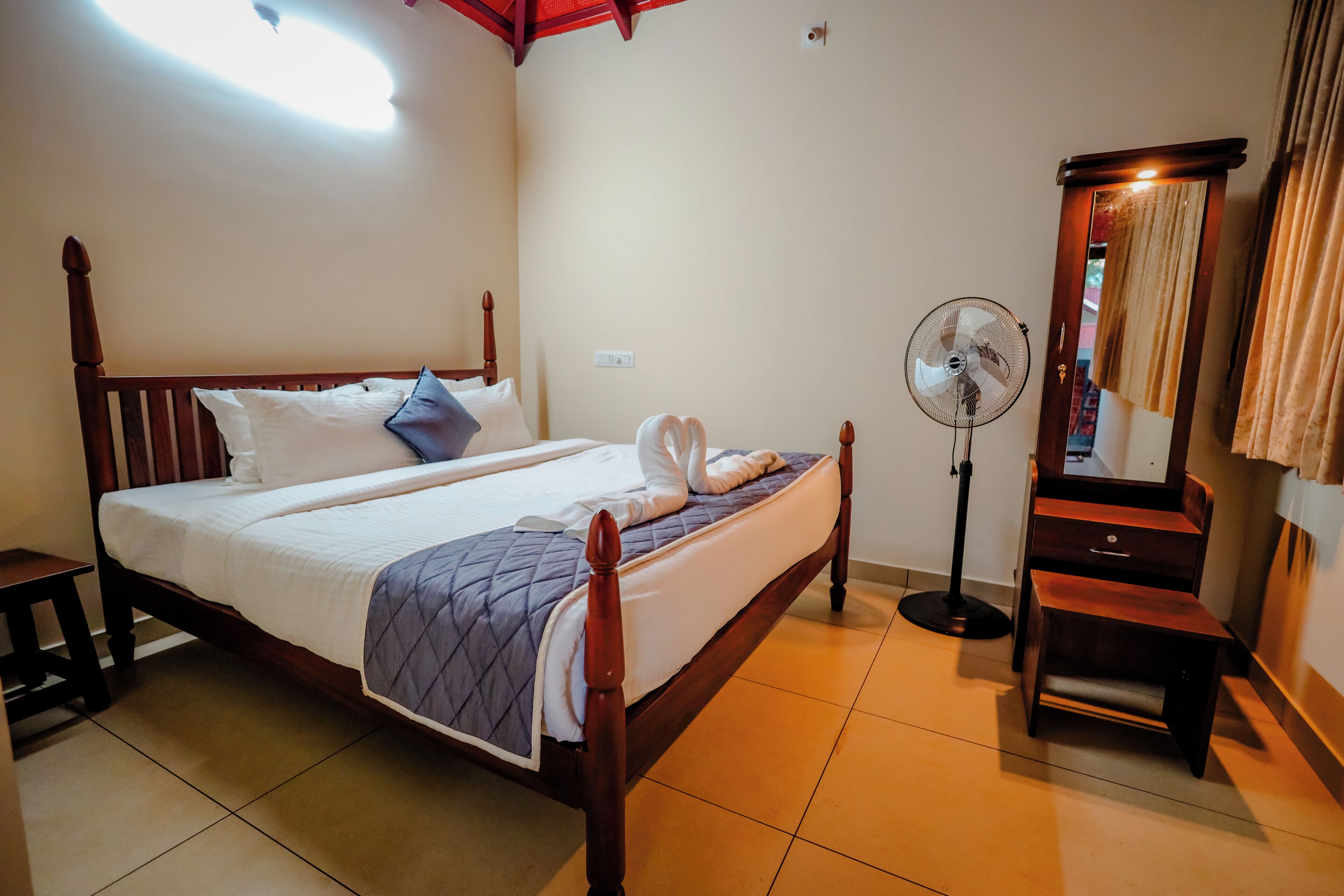 A neatly made bed with a wooden frame, a standing fan, and a mirror on the wall.