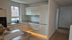 A modern kitchen with white cabinets, a wooden dining table, and a window with a view of a house. - Premium Lake Constance Base Apartment – Modern, Cozy & Well-Maintained (Rielasingen-Worblingen)