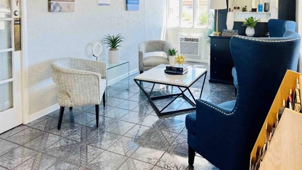 Una habitación con un sillón azul, un sillón blanco, una mesa de centro de vidrio y una mesita con una planta.