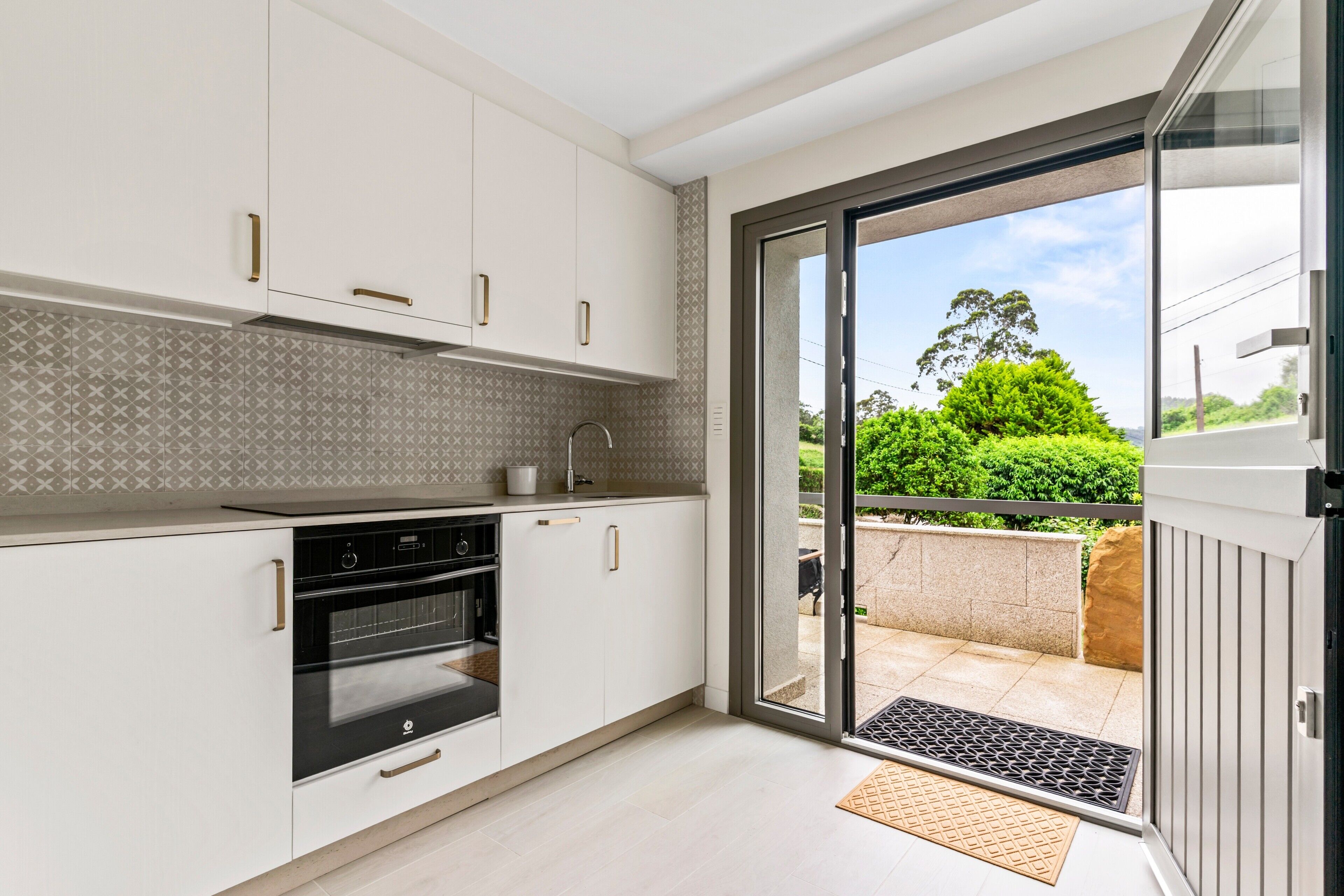 Une cuisine moderne avec des meubles blancs, un four noir et une porte-fenêtre coulissante donnant sur un balcon.