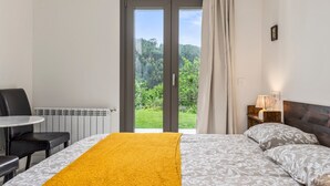Et hotelværelse med seng, bord, stol, et vindue med udsigt til grøn vegetation og en radiator.