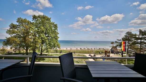 A beach view from an outdoor seating area with tables and chairs, a flag, and a tree. - Strandkoje 203 - Holiday apartment Scharbeutz directly on the beach - Strandkoje 203 for (Scharbeutz)