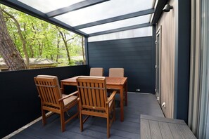 Eine überdachte Terrasse mit Tisch und Stühlen, Blick auf Bäume und ein dunkel gefasstes Gebäude.