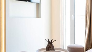 Quarto de hotel moderno com televisão de tela plana na parede, uma mesa redonda com um centro de mesa trançado, dois pufes redondos e uma grande janela com cortinas.