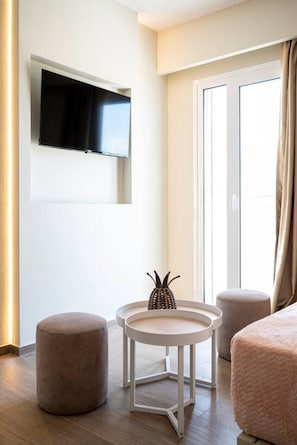 A modern hotel room with a flat-screen TV mounted on the wall, a round table with a woven centerpiece, two round ottomans, and a large window with curtains. - SolMare Deluxe Suites (Mastihari)