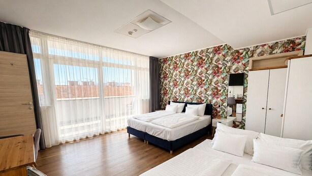 Quarto de hotel com uma cama grande, uma cama menor, um guarda-roupa, uma escrivaninha com abajur e uma janela com cortinas translúcidas.